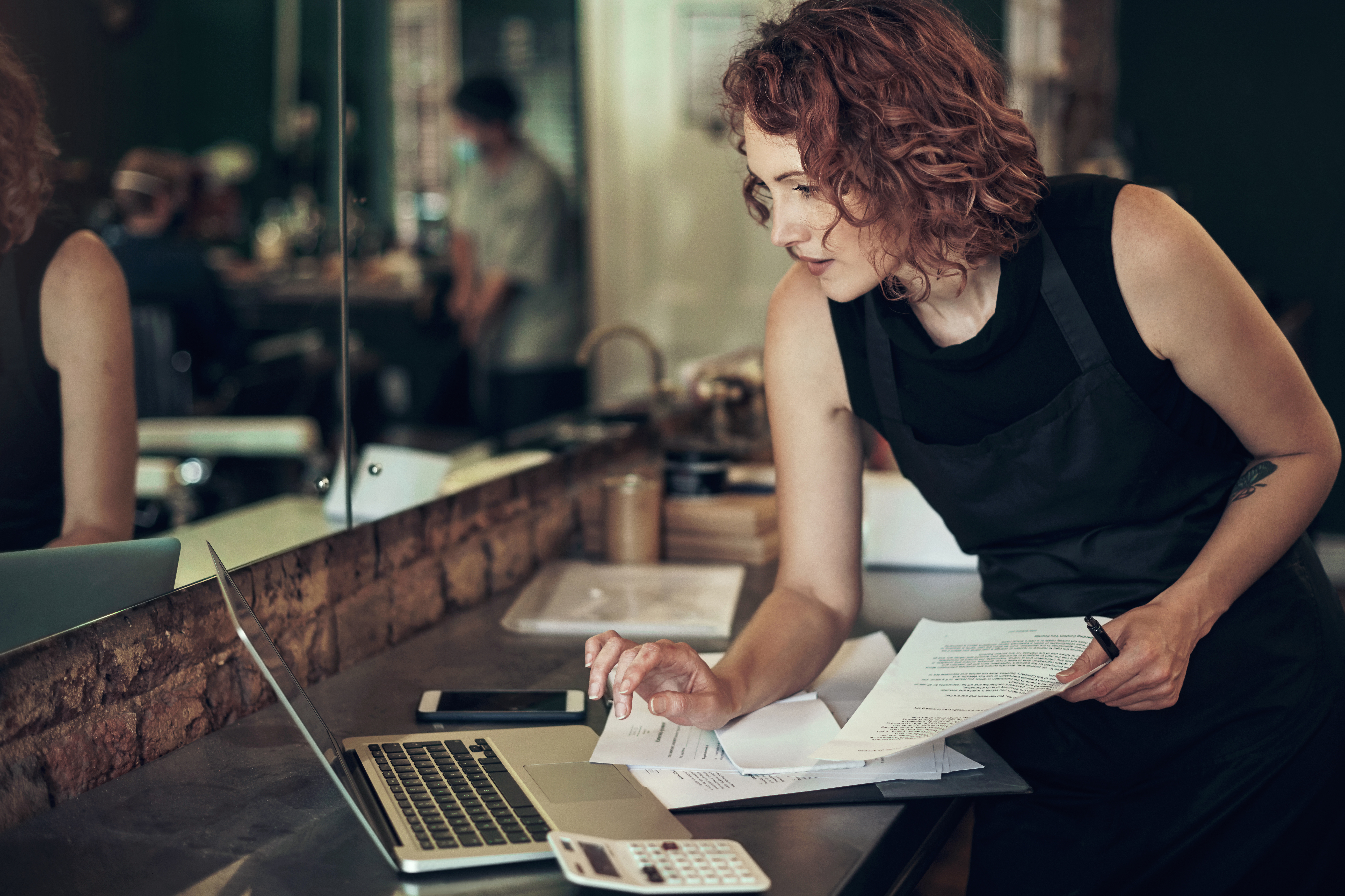 Woman holding a piece of paper consults a calculator and uses a computer. 